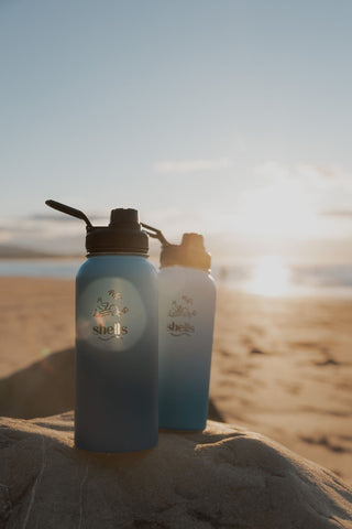 Pictured is a Shells Cafe blue water bottle on the beach with the sun hitting it